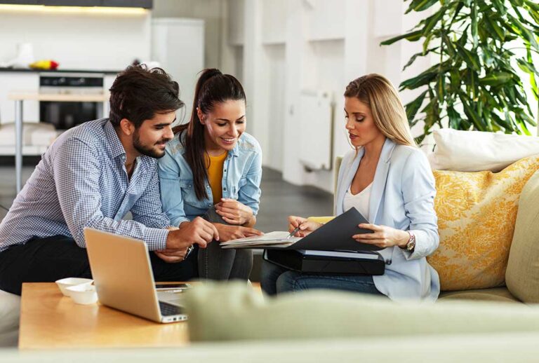 A couple and an agent discussing a home offer at a kitchen table with visible paperwork and a mountain view outside.